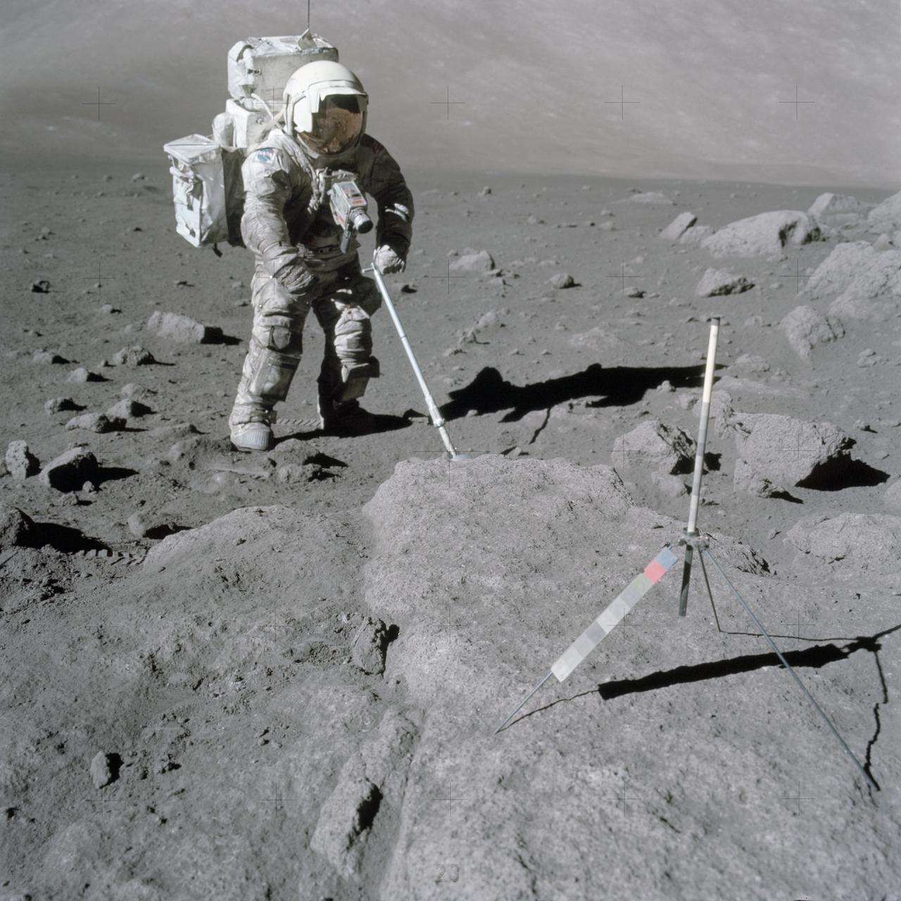 Scientist-astronaut Harrison Schmitt, Apollo 17 lunar module pilot, uses an adjustable sampling scoop to retrieve lunar samples during the second Apollo 17 extravehicular activity (EVA), at Station 5 at the Taurus-Littrow landing site. A gnomon is atop the large rock in the foreground. The gnomon is a stadia rod mounted on a tripod, and serves as an indicator of the gravitational vector and provides accurate vertical reference and calibrated length for determining size and position of objects in near-field photographs. The color scale of blue, orange and green is used to accurately determine color for photography. The rod of it is 18 inches long. The scoop Dr. Schmitt is using is 11 3/4 inches long and is attached to a tool extension which adds a potential 30 inches of length to the scoop. The pan portion, obscured in this view, has a flat bottom, flanged on both sides with a partial cover on the top. It is used to retrieve sand, dust and lunar samples too small for the tongs, another geological tool used by the astronauts. The pan and the adjusting mechanism are made of stainless steel and the handle is made of aluminum. Within the foreground of this scene, three lunar samples were taken--numbers 75060, 75075 and 75080. Astronaut Eugene A. Cernan, crew commander, was using a 60mm lens on the 70mm Hasselblad camera and type SO-368 film to take this photograph.