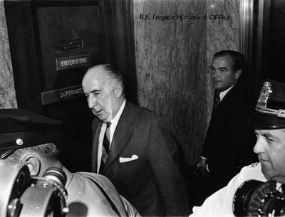 Former Attorney General John Mitchell enters the Senate caucus room to testify, 1973. Archival image available at modest resolution.